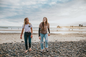 Two blonde girls, wearing jeans and T-shirts. Also wearing vegan Noz sunscreen walking on the beach. The sky is blue with clouds.